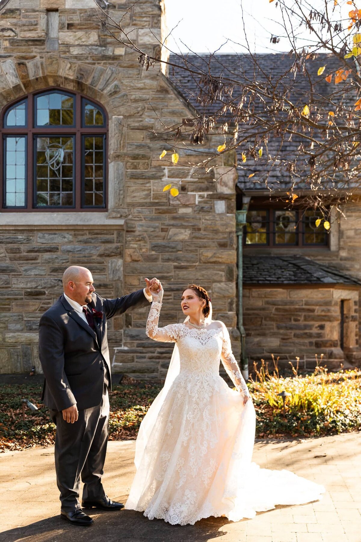 Beneath the golden glow of autumn sunlight, the groom lifts the bride’s hand for a twirl outside a historic stone chapel. Her long-sleeved lace wedding dress flows elegantly around her, and his dark suit and deep red boutonniere add a classic contrast against the rustic charm of the church’s stained glass windows
