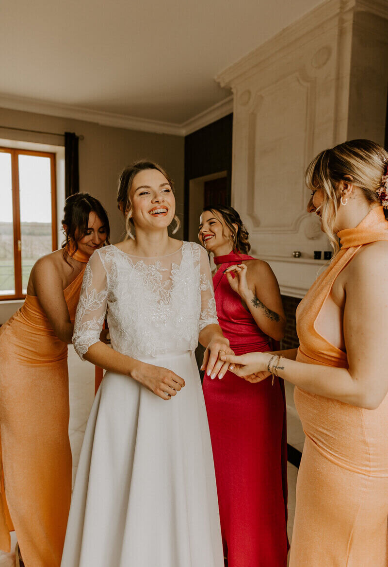 Mariée, riant, entourée de ses demoiselles d'honneur, habillées couleur pêche et corail devant une cheminée lors d'un shooting photo mariage.