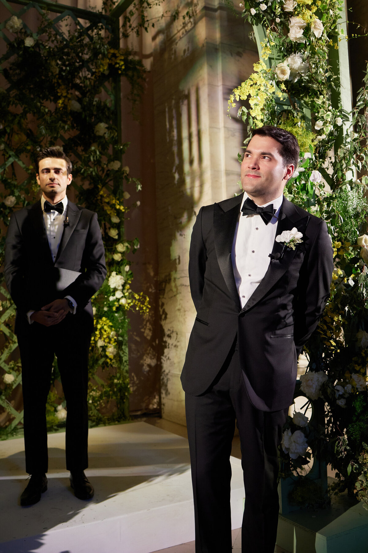 The photo was taken at the iconic New York Public Library in New York City by photographer Shawn Connell. It captures two men in tuxedos within a well-lit setting adorned with greenery and flowers. The man in the front charmingly looks to the side with a smile, his hands elegantly placed behind his back. Meanwhile, the other man stands in the background, gazing forward with a neutral expression, adding an air of sophistication to the scene. Additionally, Aaron Navak Films provided videography services for this event, capturing the elegant atmosphere on film.