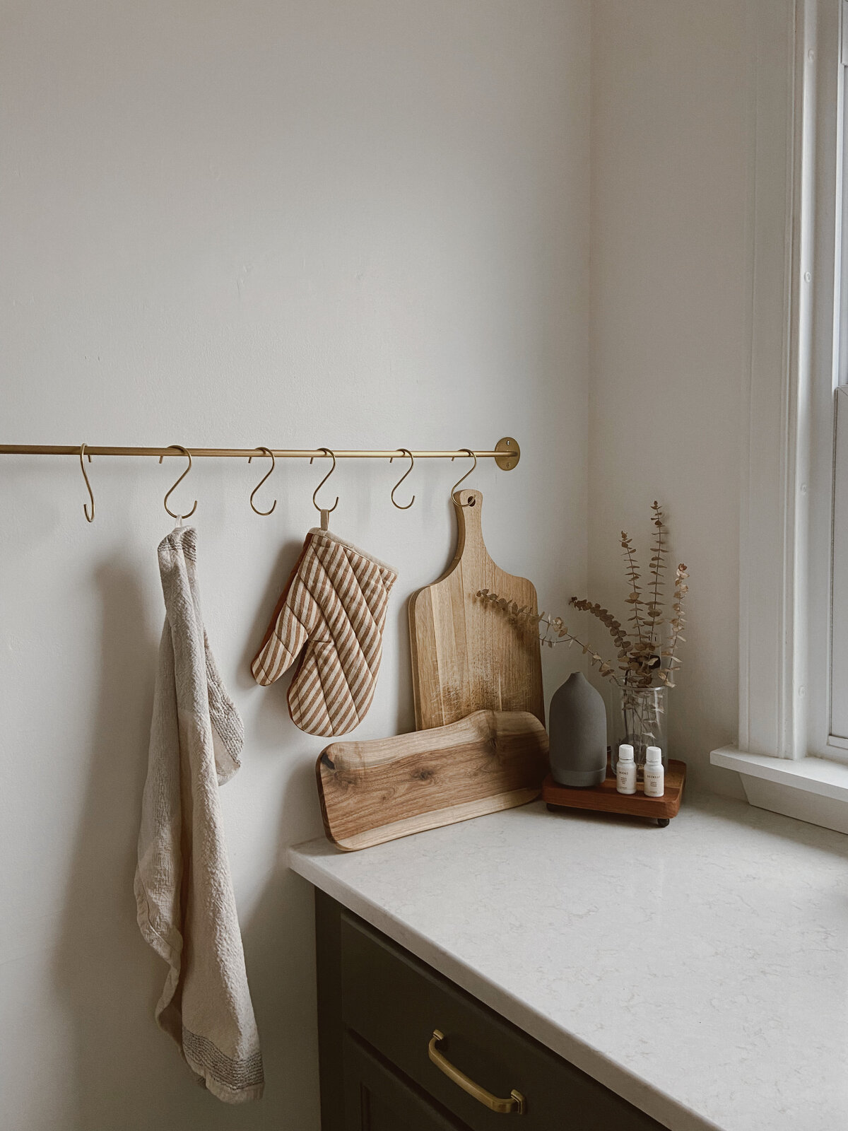 A beautifully designed bakery kitchen featuring a dark wooden counter with a white marble counter top. A beige tea towel and striped brown-and-white oven gloves hang on hooks, while wooden breadboards lean against the wall. A wooden tray in the corner holds a glass vase with dried eucalyptus and an oil diffuser, creating a warm and inviting workspace for baking.