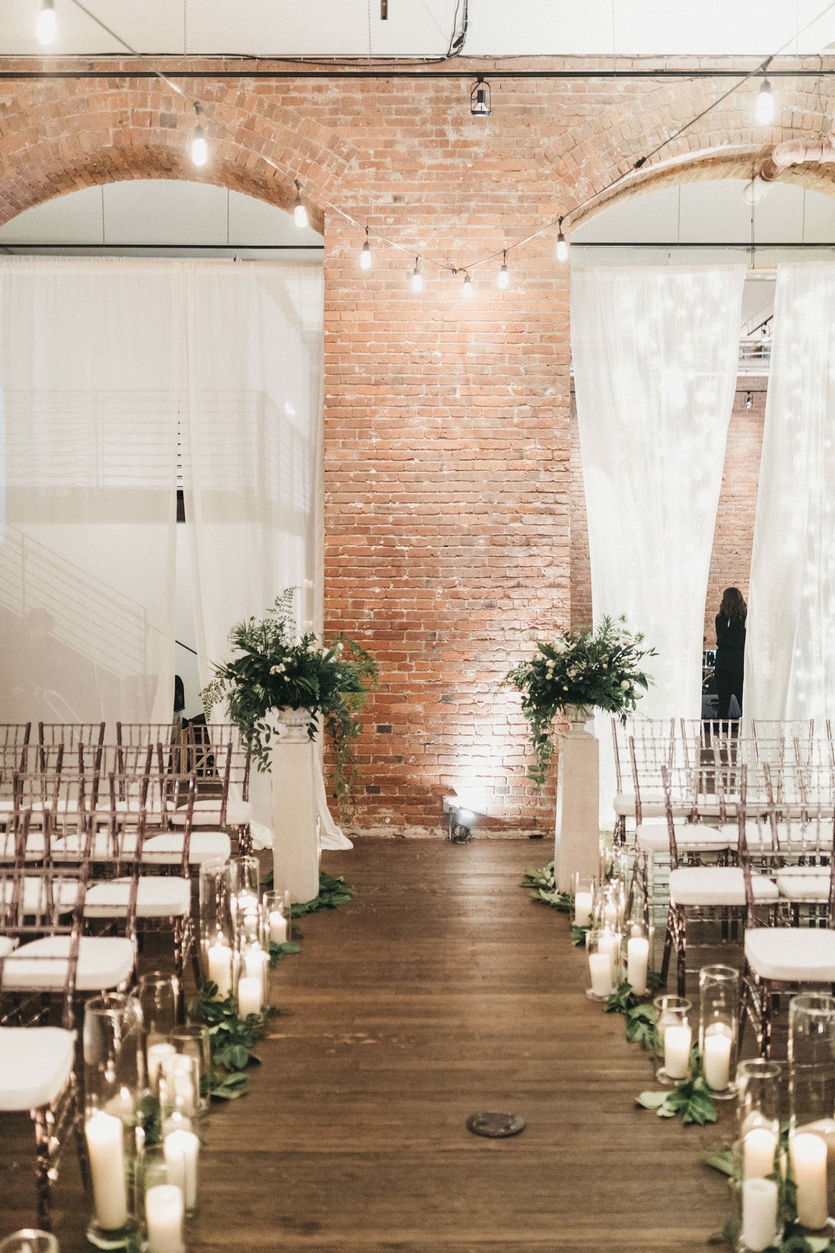 wedding aisle with lots of hurricane candles, lined with greenery, and tall greenery arrangements at aisle entrance