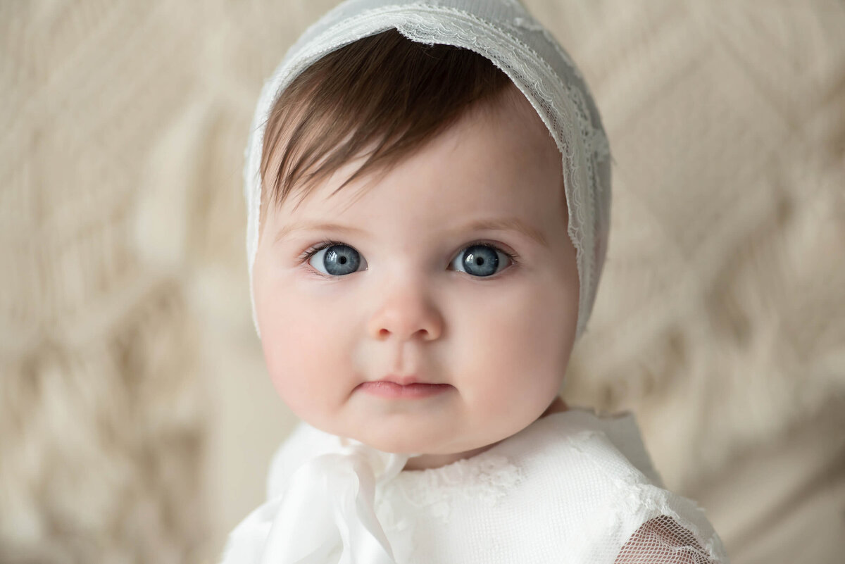 Baby girl wearing a vintage bonnet and white christening dress in front of a beige background for her kids photos in Burlington, ON