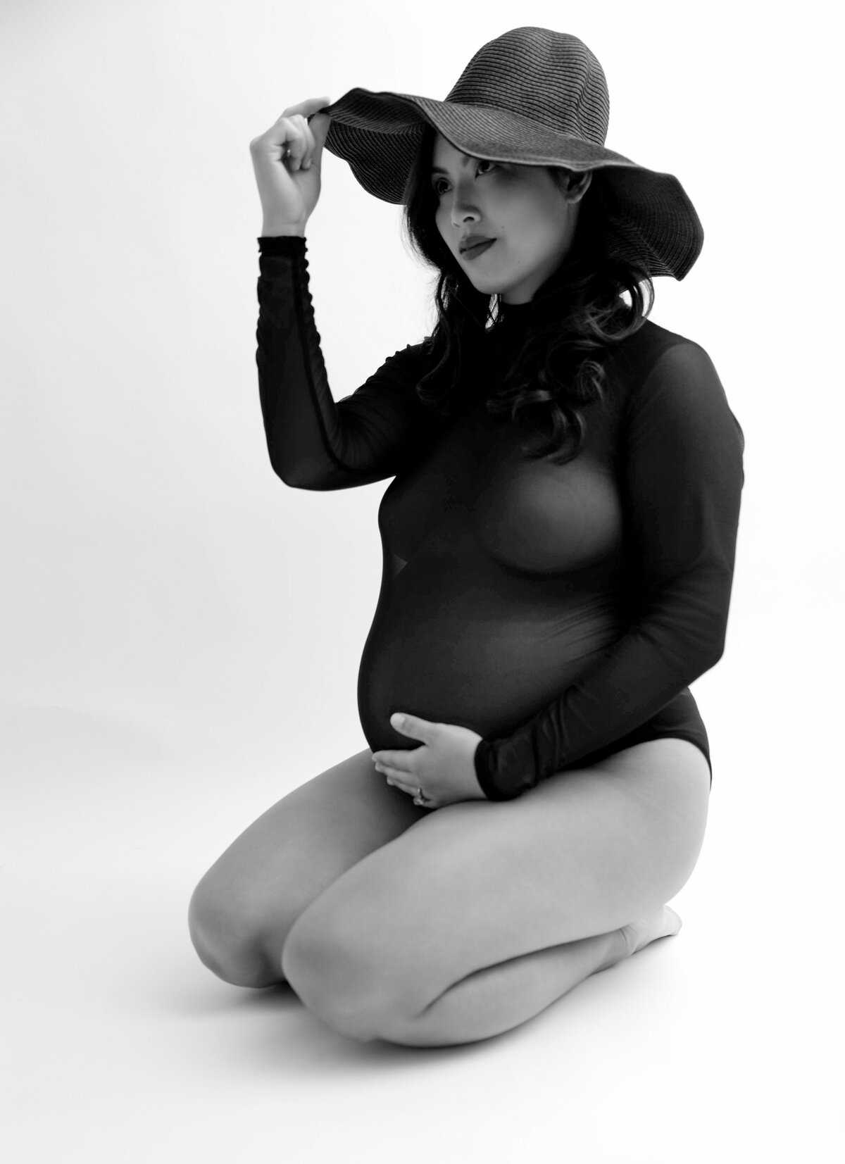 Pregnancy photo of mother wearing a black bodysuit with one hand posed under belly and the other hand slightly touching the rim of a black summer hat.