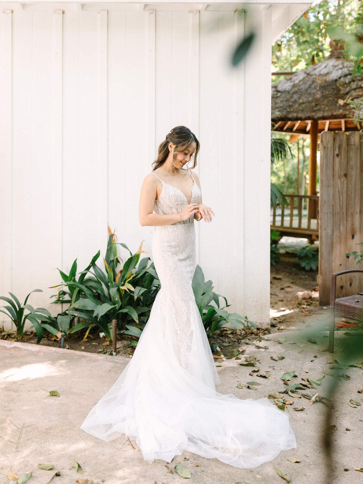 Bride in a lace wedding dress stands outside near plants, smiling gently.