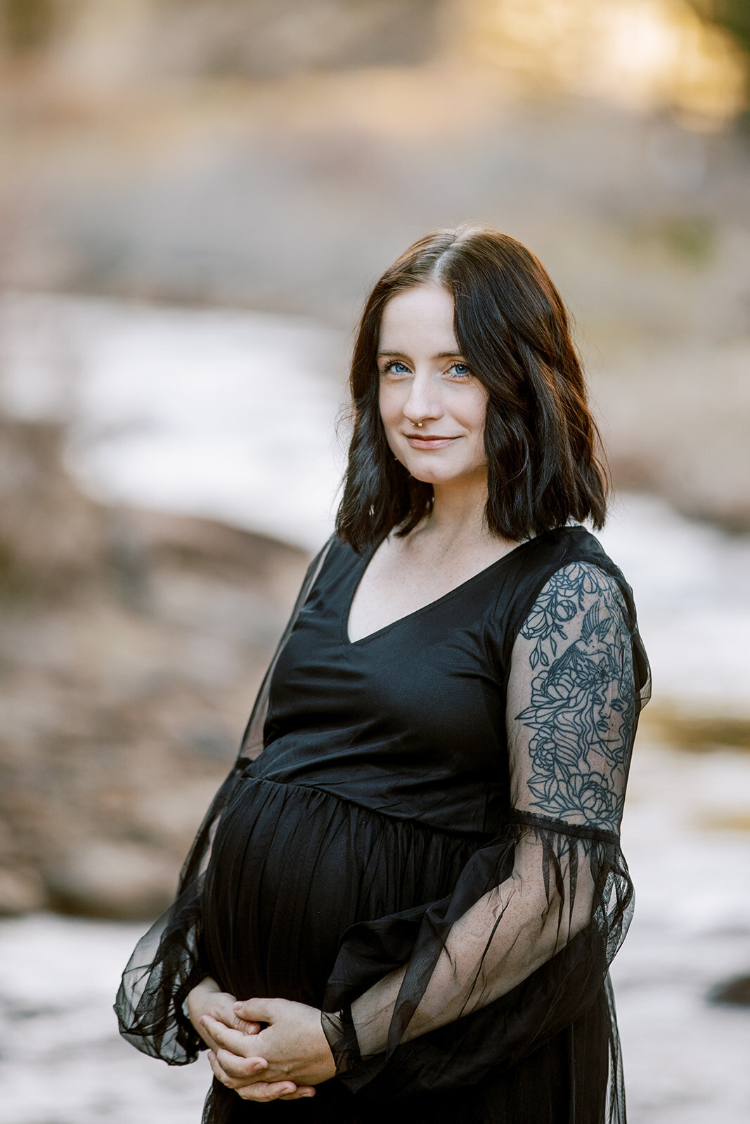 pregnant woman in a black dress and tattooed arm holds her big belly near a colorado river