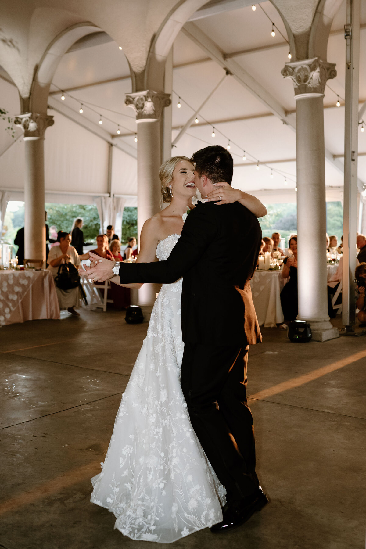 Bride and groom share their first dance under a white tent with glowing string lights.
