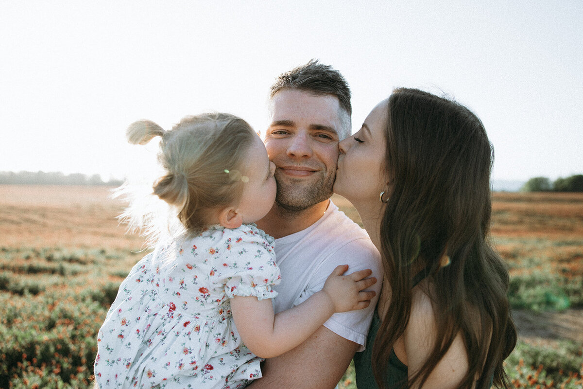 Mother and daughter give dad kisses on either side of his cheeks during sunset near a clover field in Oregon. 