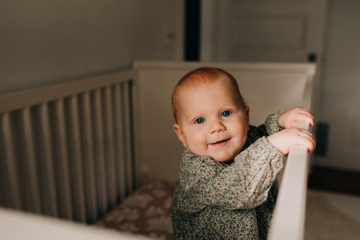 A toddler with blue eyes and red hair stands in her crib wearing a green floral outfit