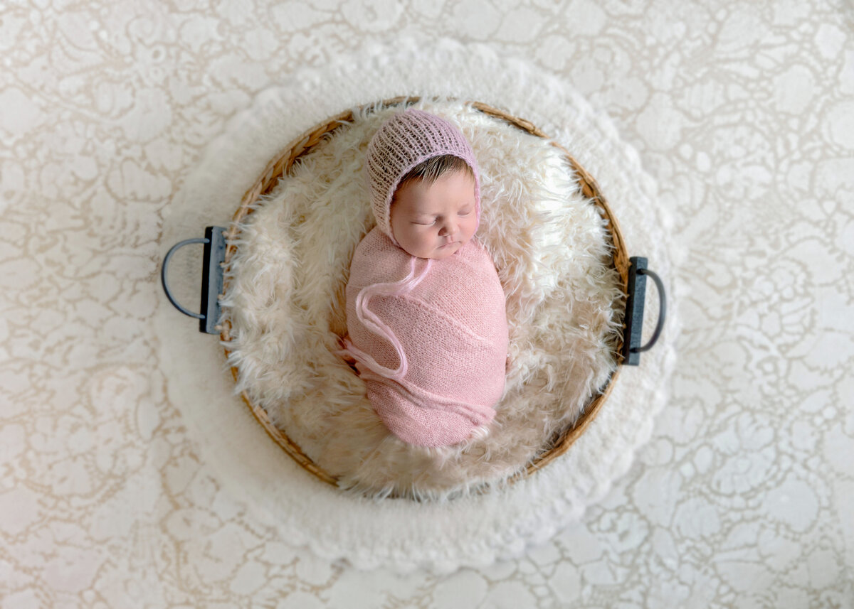 A newborn baby in a pink wrap  and wearing a white bonnet, in basket  that is on top of a  cream colored lace  backdrop.