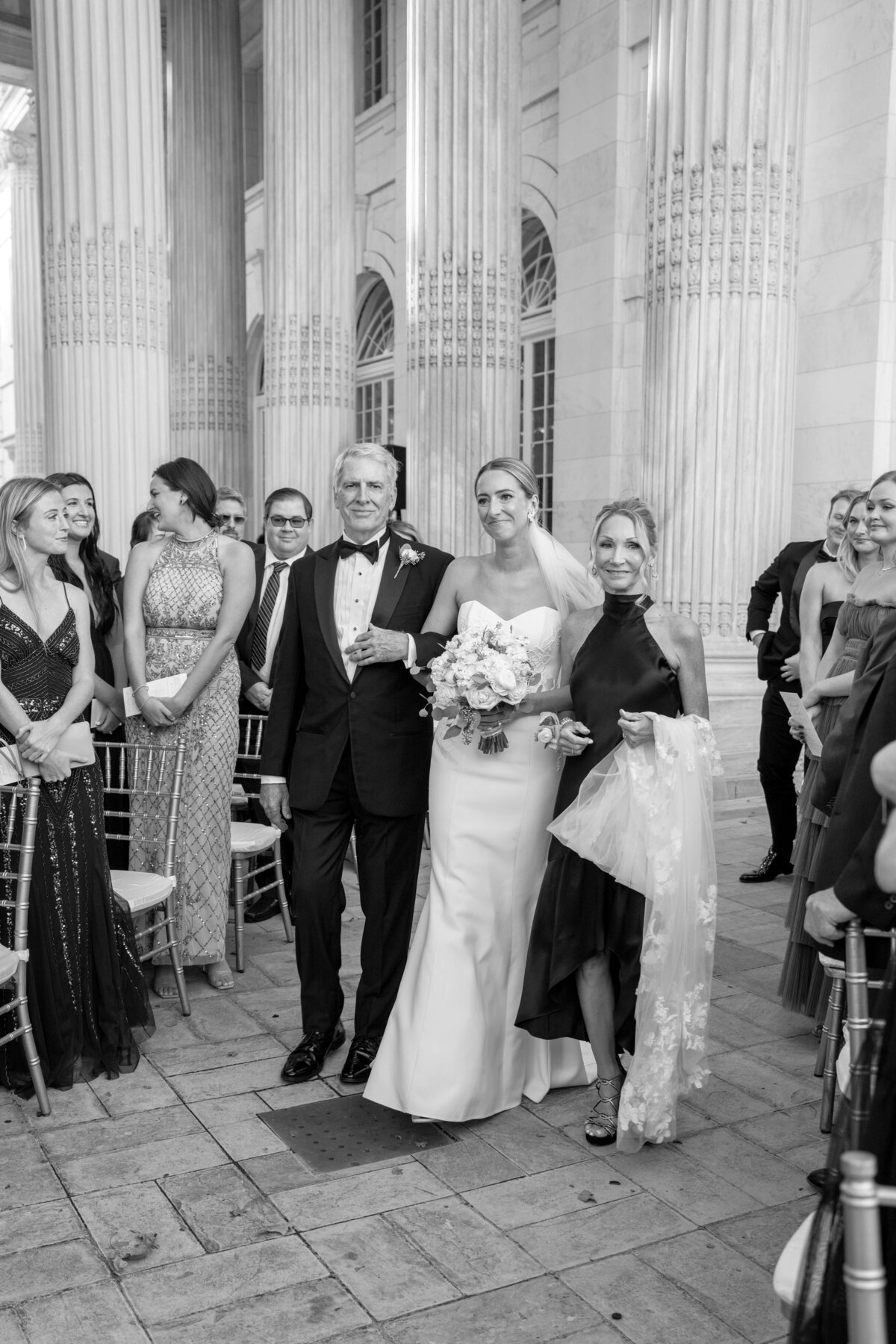 A bride in a white dress, holding a bouquet, walks down the aisle accompanied by two people. They are surrounded by smiling guests. The setting features elegant columns and chairs, suggesting a formal indoor wedding. The image is in black and white.