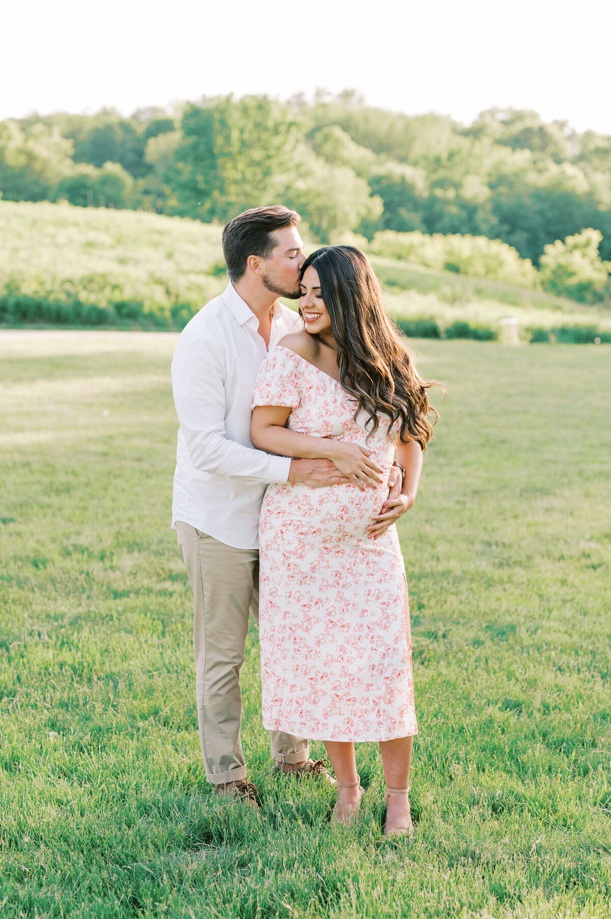 Mom and dad standing together in field with hands on mom's stomach during maternity session
