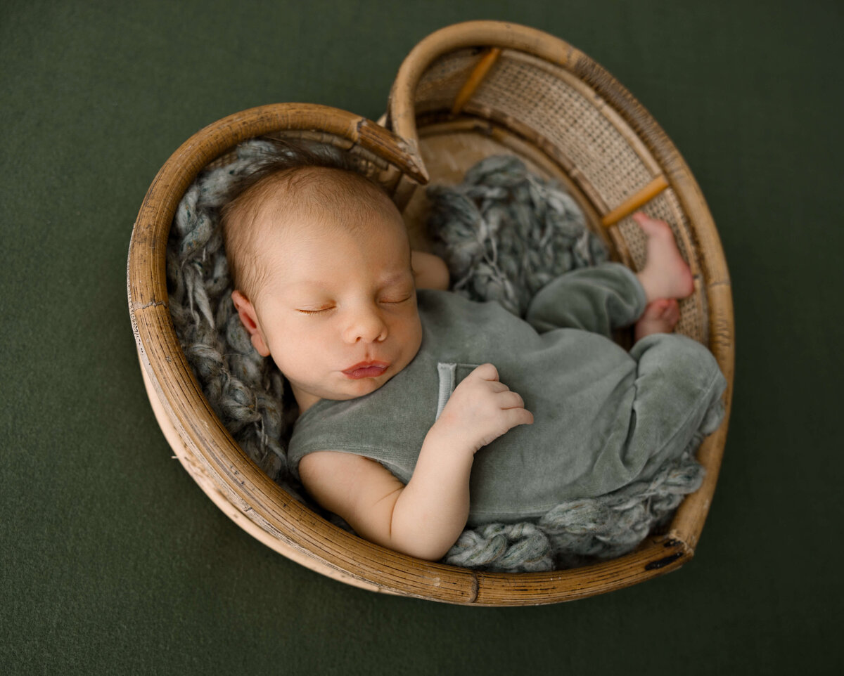 Little newborn baby boy wearing green in a heart shaped basket and sleeping