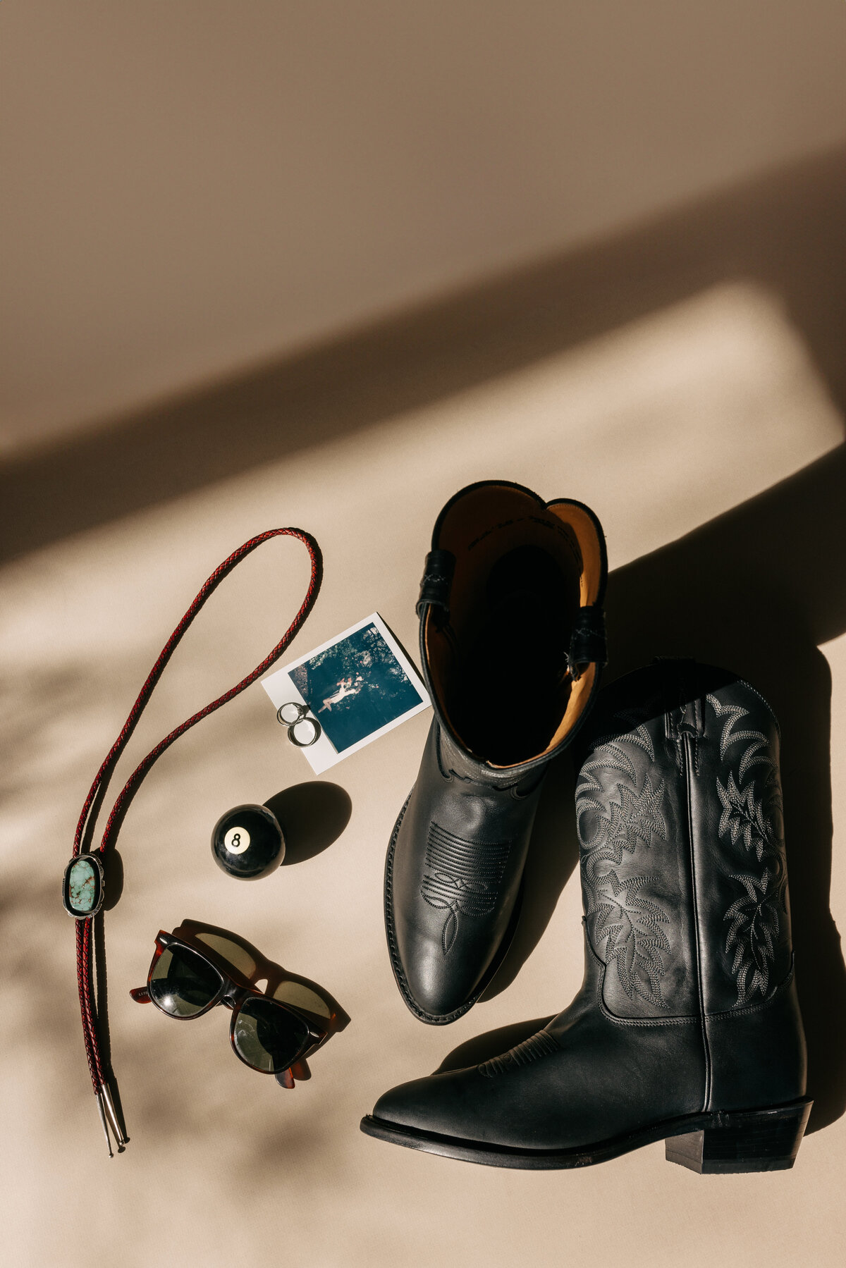 jade bolo tie sunglasses 8 ball polaroid wedding rings and black cowboy boots laying in the sunlight for a wedding detail shot