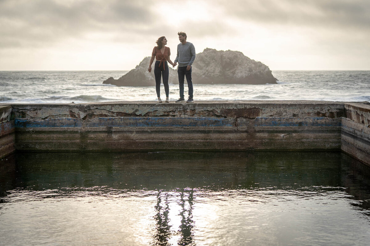 couple standing on the edge of sutro baths with a reflection of them in the water at sunset