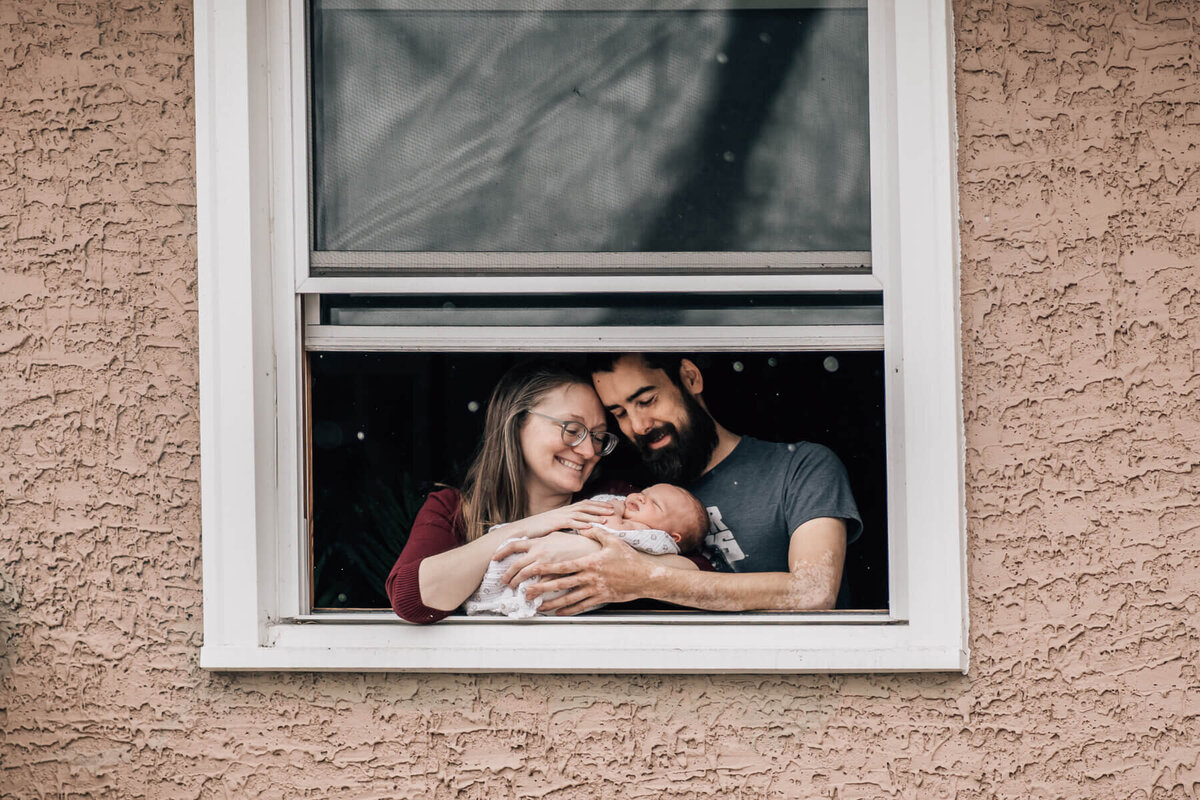 New parents cradle their newborn child in the window of their home, captured by Minneapolis newborn photographer, Kate Simpson.