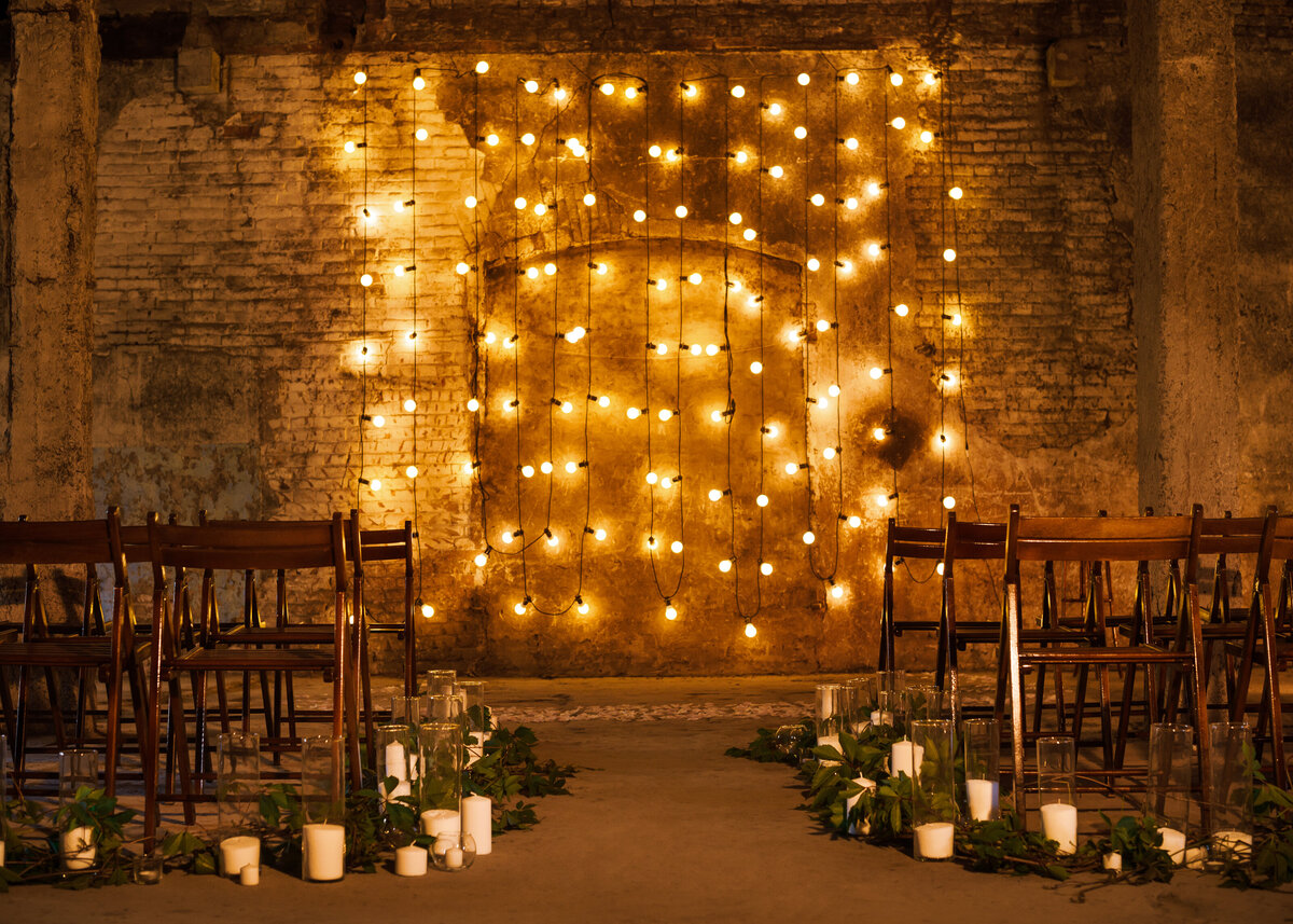 Wedding ceremony backdrop of festoon lighting hangs at the end of the aisle lined with candles and garlands in a brick room