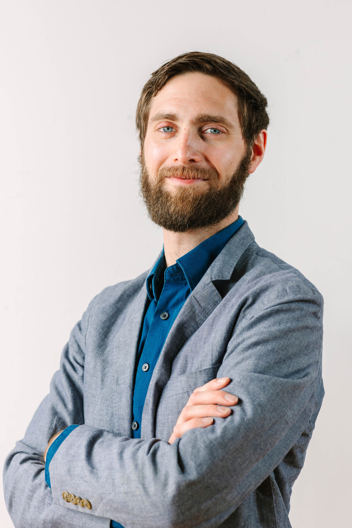 A confident man with a well-groomed beard and short brown hair stands with his arms crossed against a plain white background. He wears a gray blazer over a deep blue button-up shirt, giving a polished and approachable look.