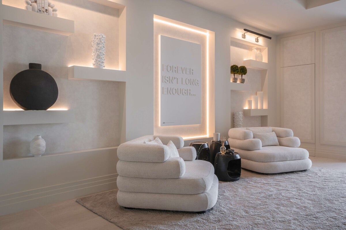 A spacious, modern entry area at the foot of the stairs, with a focus on light, neutral tones. There are two plush, light-colored armchairs and a soft, textured rug. Built-in shelves with decorative items and a wall with the illuminated text 'FOREVER ISN'T LONG ENOUGH' add a sleek coolness to the space. In the background, a dining area with a large table and pendant lights is visible through glass doors.