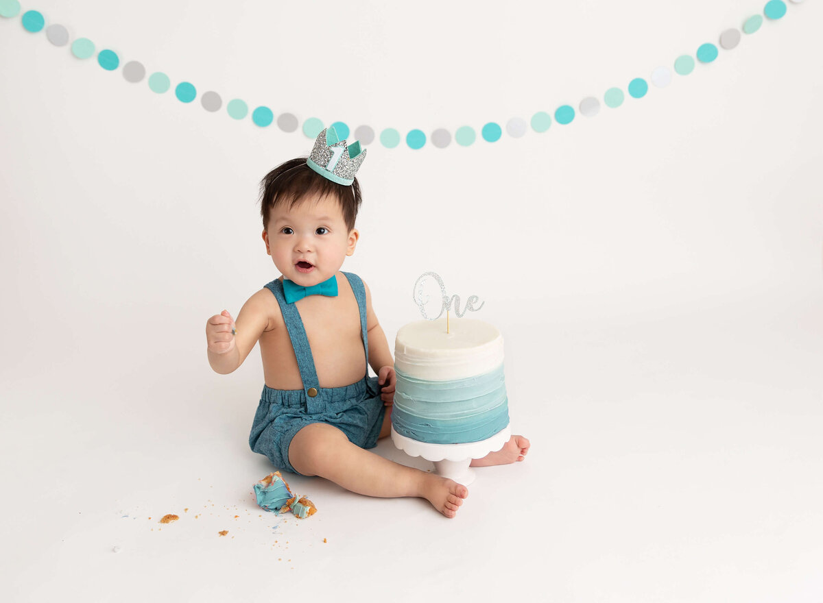 Cake smash photography in New York City featuring a baby boy enjoying their first birthday celebration with a pastel-colored cake and mint banner. Capturing joyful moments for parents seeking memorable photoshoot in NYC