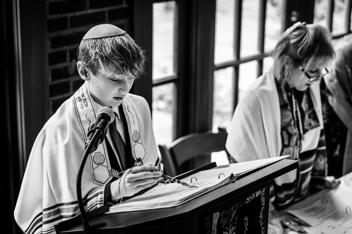 A boy wearing a traditional Jewish shawl and head covering uses a Yad to read at the podium