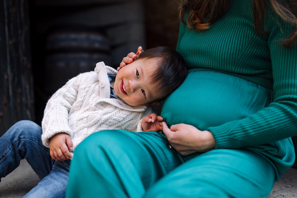 3-year-old boy sitting with pregnant mom in green dress and leaning on her baby bump