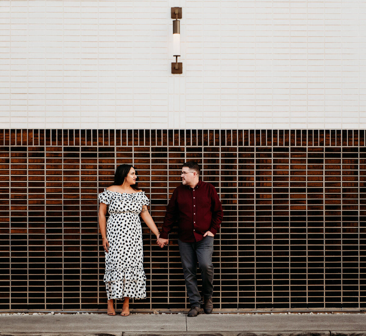 Couples Photography, Woman in black and white dress holding hands with man in maroon button down shirt, looking at each other. They are on South Congress Street against a light brown and white wall.