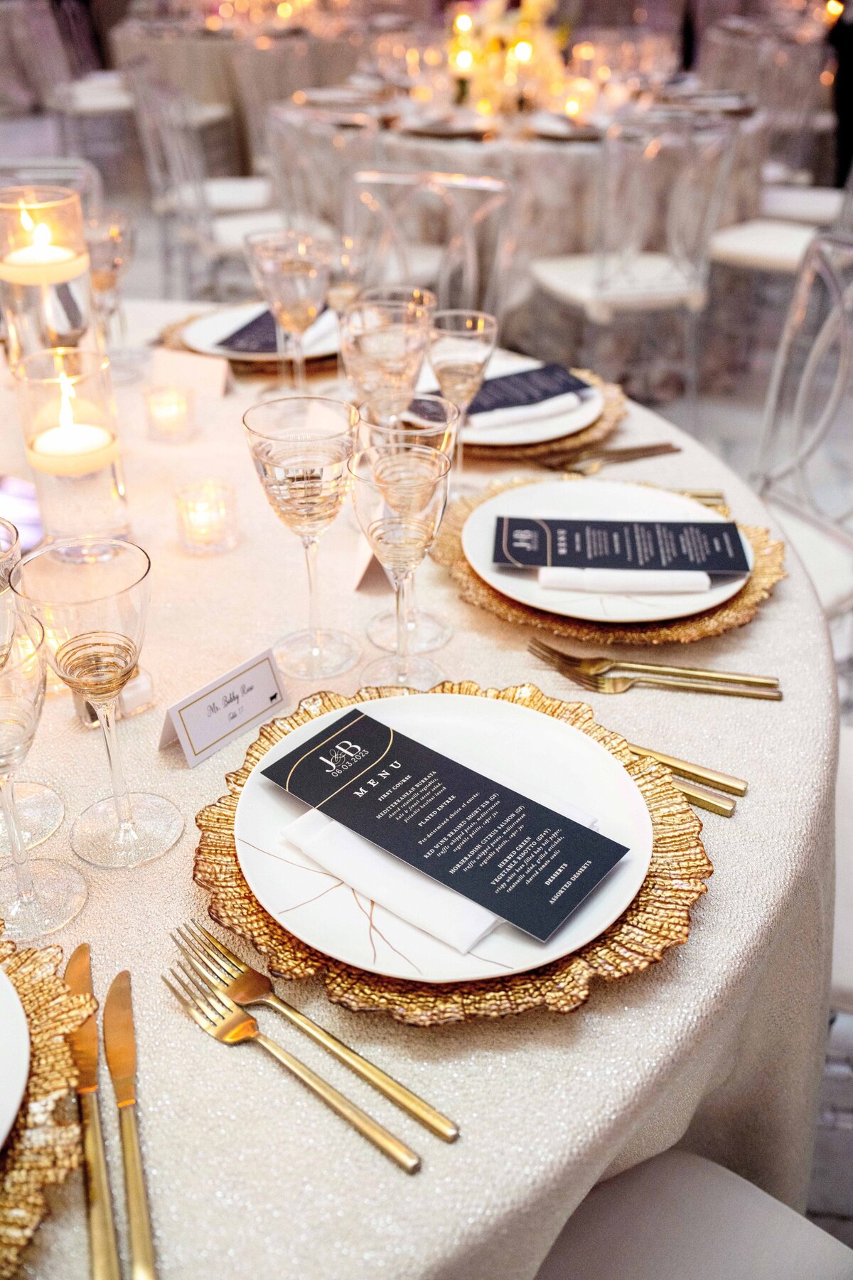 Elegant table setting with gold-rimmed plates, crystal glasses, and gold cutlery on a white tablecloth. Black menu cards rest on each plate. Lit candles and floral arrangements create a warm, inviting atmosphere.