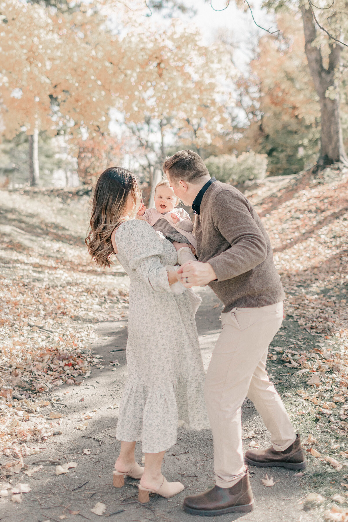 Mother and father smile at daughter while dancing together during a fall family session celebrating their daughter’s first birthday in Kansas City.