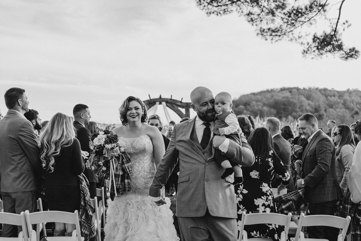 A wedding couple walking back up the aisle as one holds a baby 