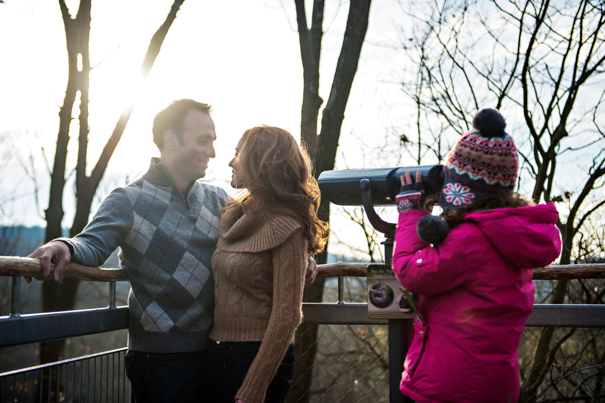 A little girl looks through a telescope at an engaged couple in morris arboretum.