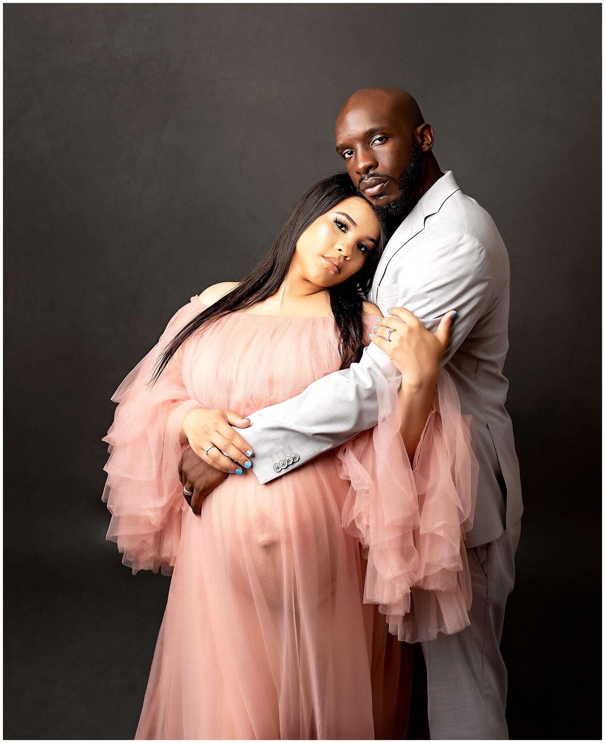A couple in a maternity photoshoot, standing close together and cradling the pregnant belly. They both are looking at the camera and just relaxing into each others arms.