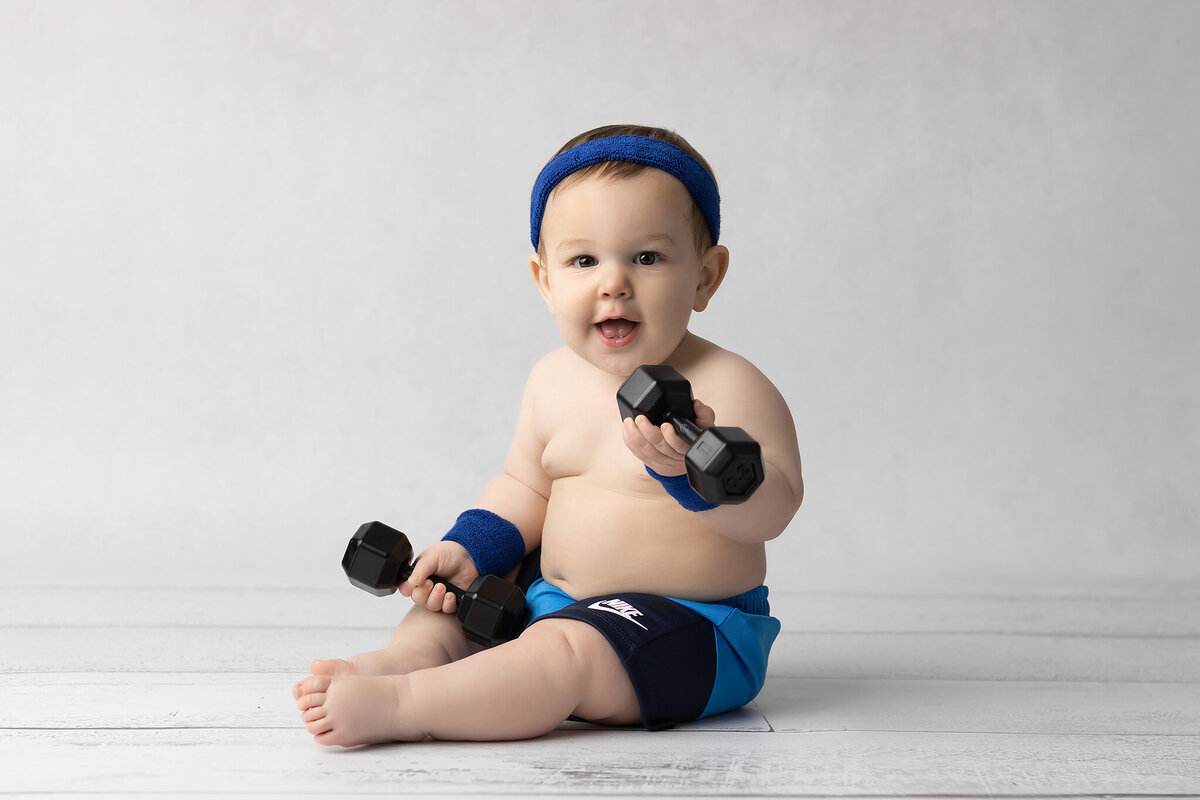 Baby boy in blue workout gear lifting toy dumbbells while sitting on a white wood floor with a light background, captured by a DFW milestone photographer.