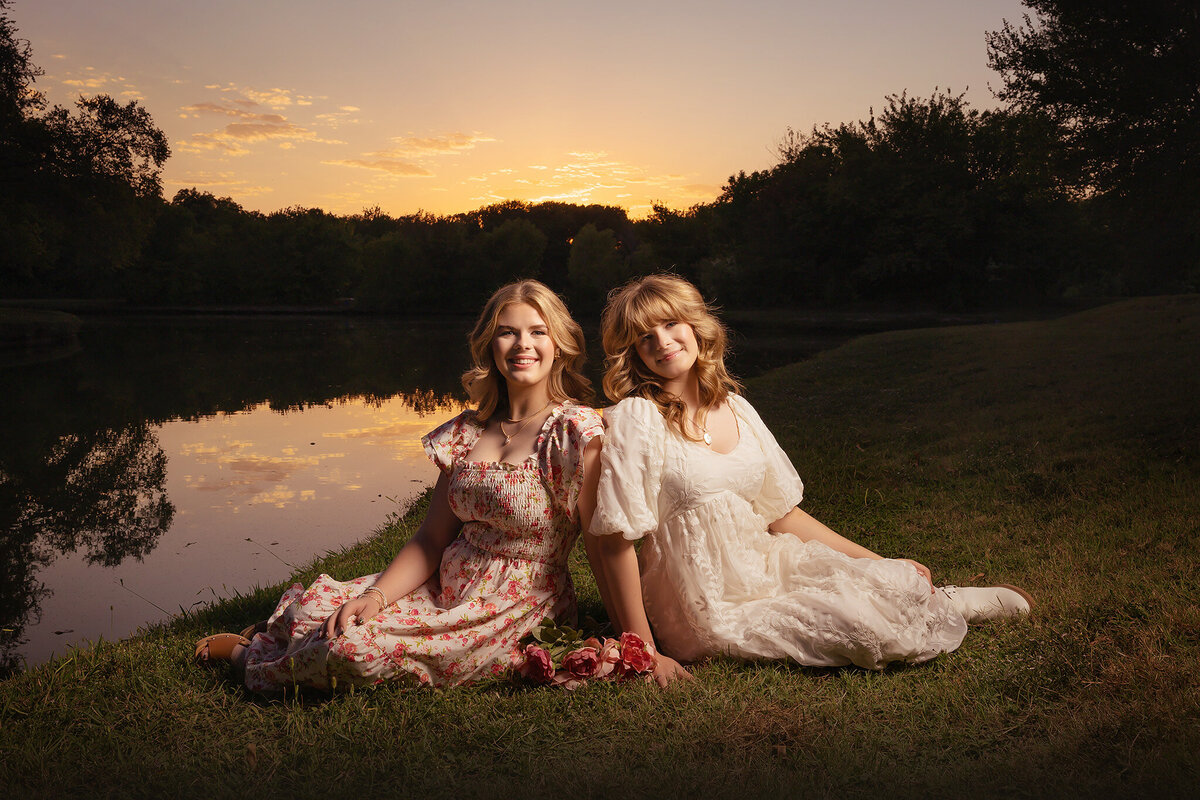 Dallas senior photographers capture two young women sitting side by side, smiling at sunset, wearing floral and white dresses, with the reflection of the sky and trees in the water behind them.