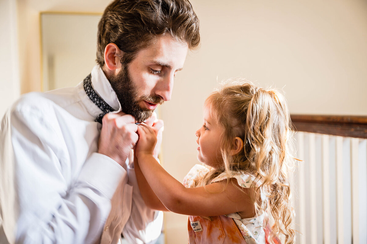 On his wedding day, a groom ties a bowtie around his neck while his daughter attempts to help.