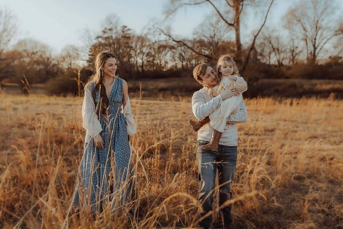 A family of three in a field, with tall grass and trees. A woman in a blue dress stands smiling; a man holds a child in his arms. The background shows a clear sky and bare trees.