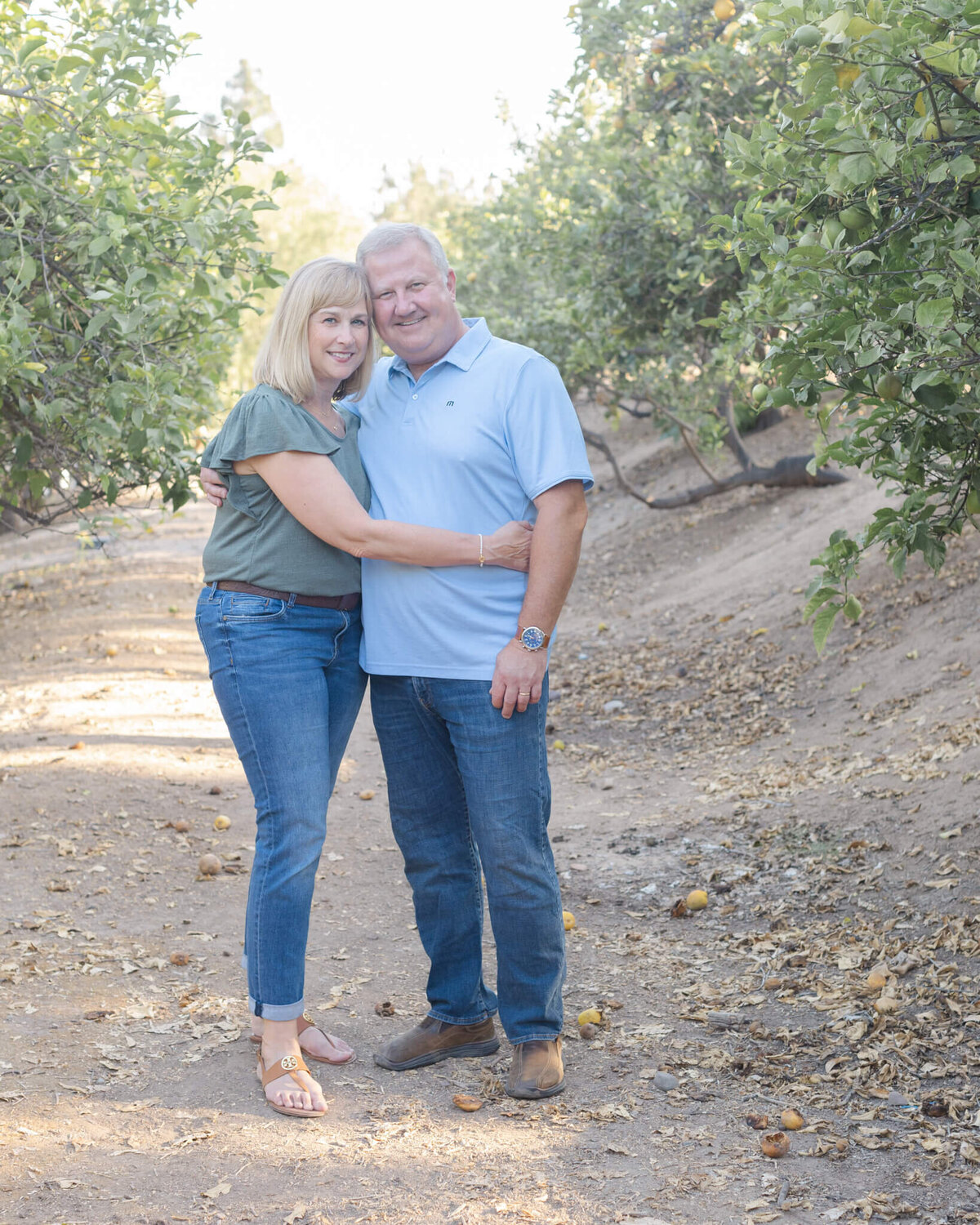A husband and wife with their arms around each other, hugging on a dirt path surrounded by green trees.