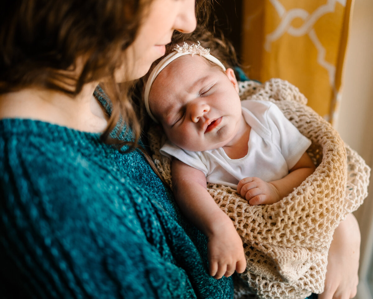 The essence of motherhood with top Albuquerque maternity photographers. The image features a woman in a blue sackcloth holding a newborn baby girl wrapped in a beige blanket and adorned with a matching headband, both peacefully resting with their eyes closed.