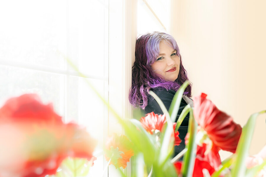 Senior portrait of young woman with frosted purple hair leaning against a white wall indoors with red flowers in the foreground. 