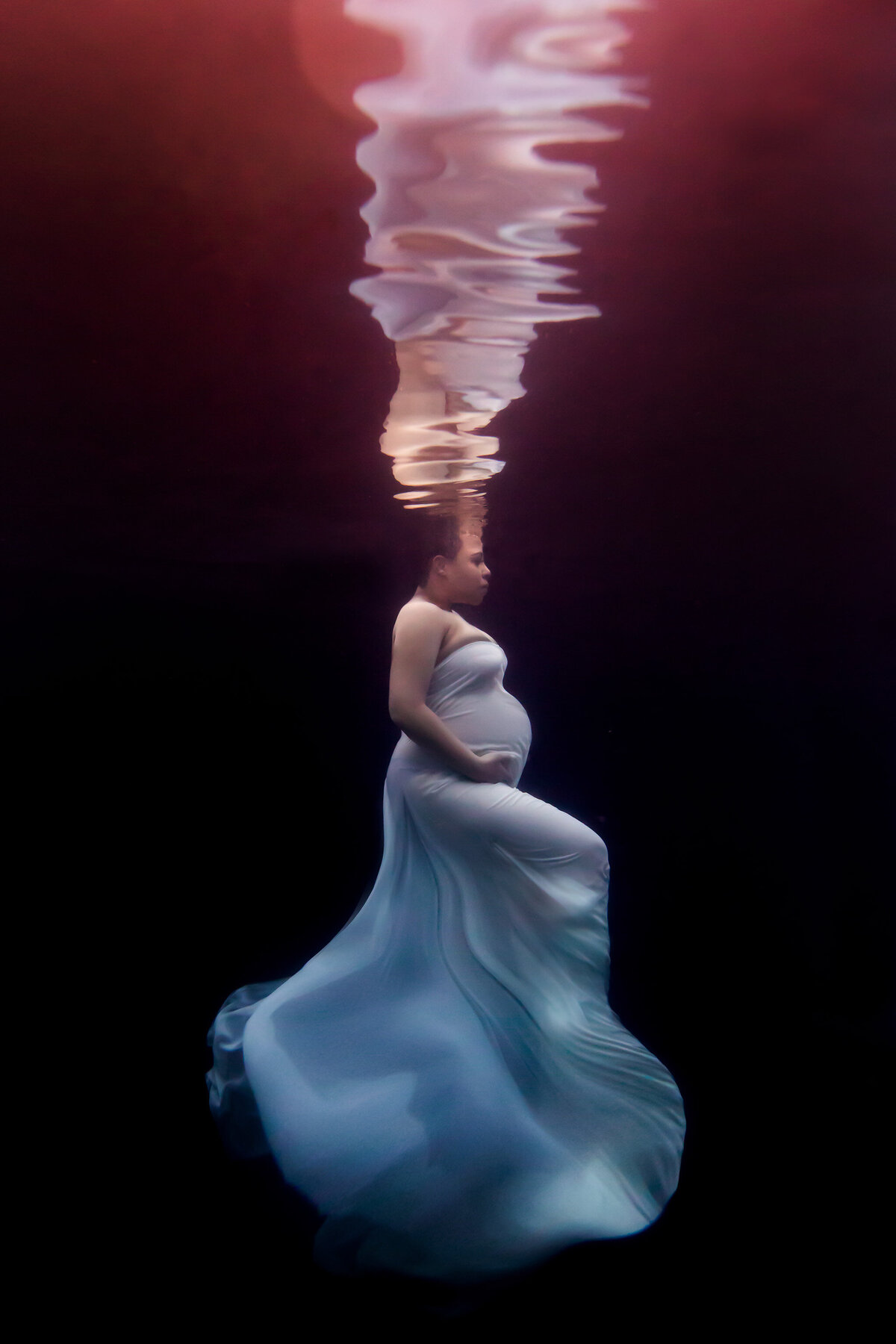 A stunning underwater pregnancy photo captured by a Dallas underwater pregnancy photographer. The woman, gracefully suspended in dark water, wears a flowing white gown that swirls like a gentle wave around her body. The soft red glow from above reflects off the water, creating a mesmerizing visual effect. This elegant underwater maternity photoshoot beautifully illustrates tranquility and strength.