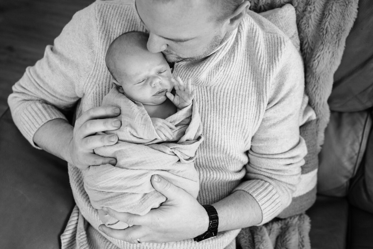 A black and white close-up of a father holding his sleeping newborn swaddled in a blanket, with the father's head gently resting against the baby's.