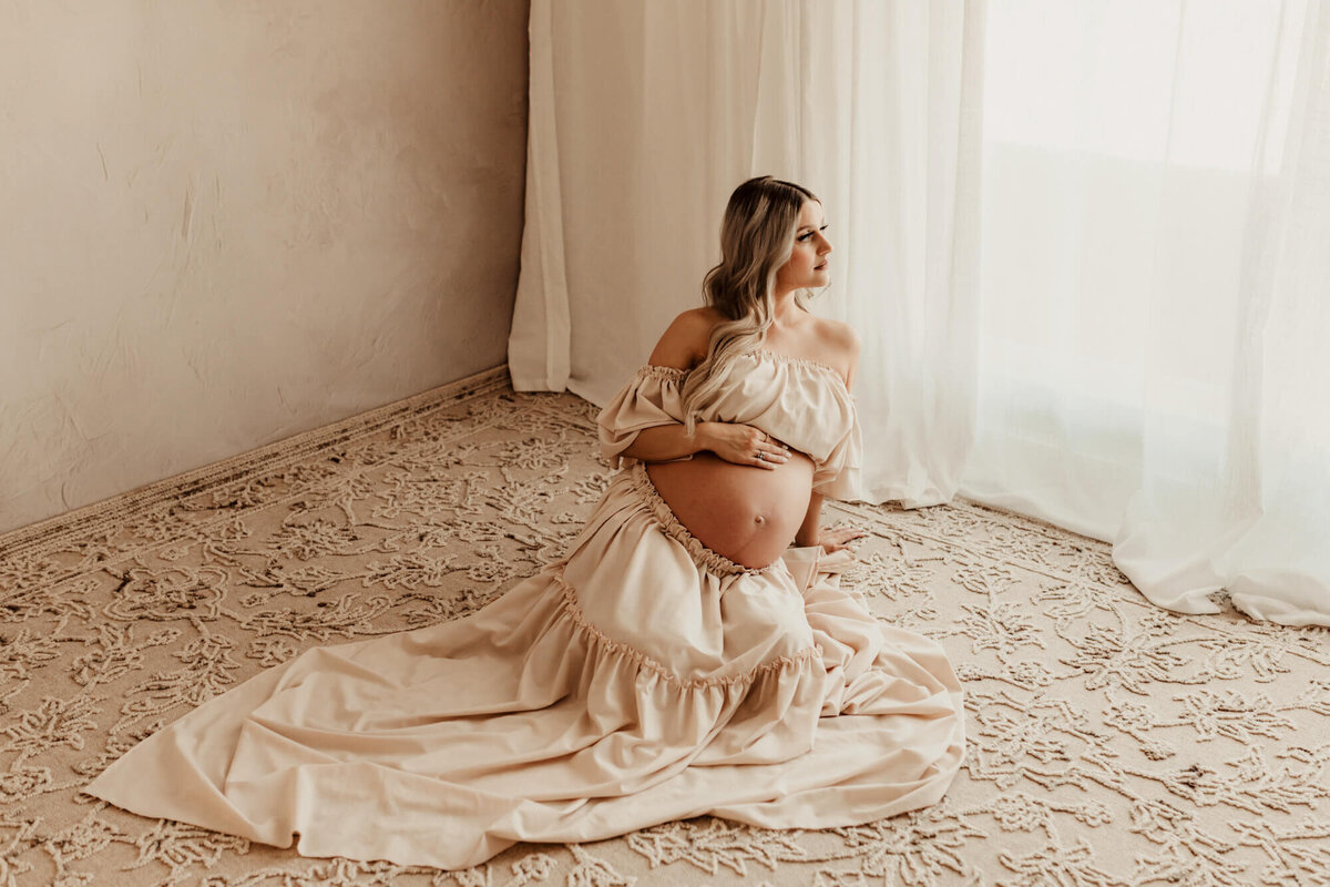 Pregnancy portrait of a woman wearing a cream colored dress sitting on a floral rug.