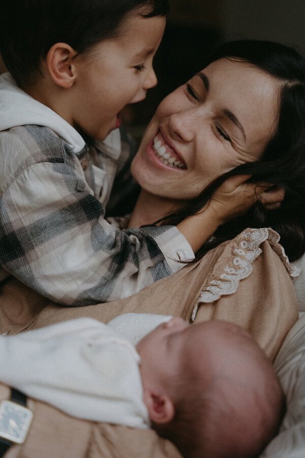 La mère éclate de rire sous les chatouilles de son fils, le bébé blotti contre elle. Shooting photo famille par Kamescop.