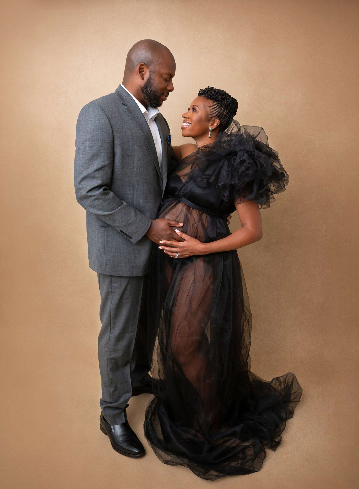 An expecting couple with hands on the bump touch foreheads while standing in a studio in a black dress after meeting a NYC maternity photographer for pregnancy pictures