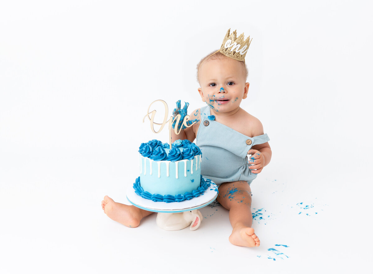 Baby boy in blue romper sits with blue cake between his legs for first birthday cake smash photoshoot. Baby has blue icing on his hands and face and is smiling at the camera.