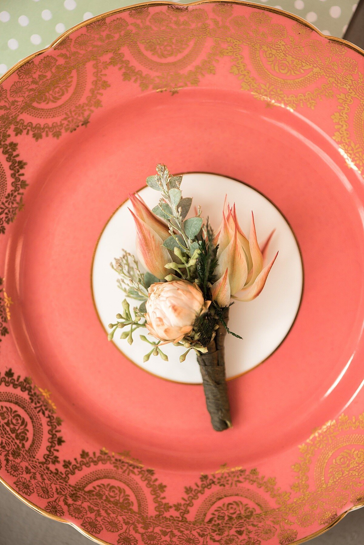 Groom's blush and peach boutonniere with a protea and tea rose on a coral vintage china dessert plate decorated with gold filagree