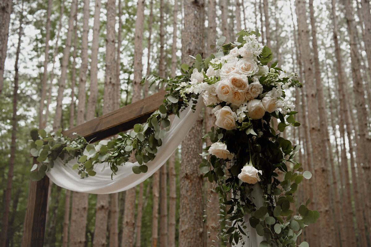 a wooden wedding arch with white drapery, roses, and greenery standing in a forest