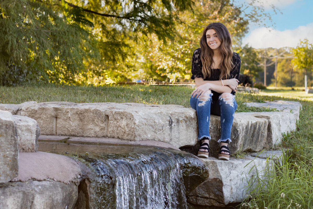 A high school senior girl sits on a stone ledge near a small waterfall, surrounded by lush greenery. She wears a stylish black lace top and ripped jeans, smiling warmly as sunlight filters through the trees. This serene outdoor setting provides a perfect backdrop for senior portrait photography.