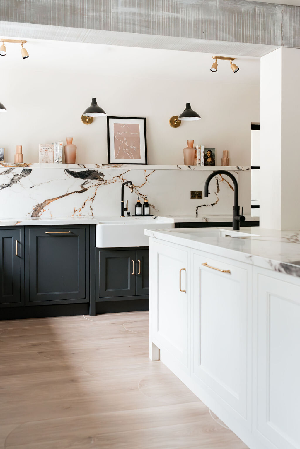 A modern kitchen with dark lower cabinets, a white marble backsplash with black and gold veining, a farmhouse sink, and minimalist open shelving holding books and decor.  A center island has matching marble counters, paired with white lower cabinets.