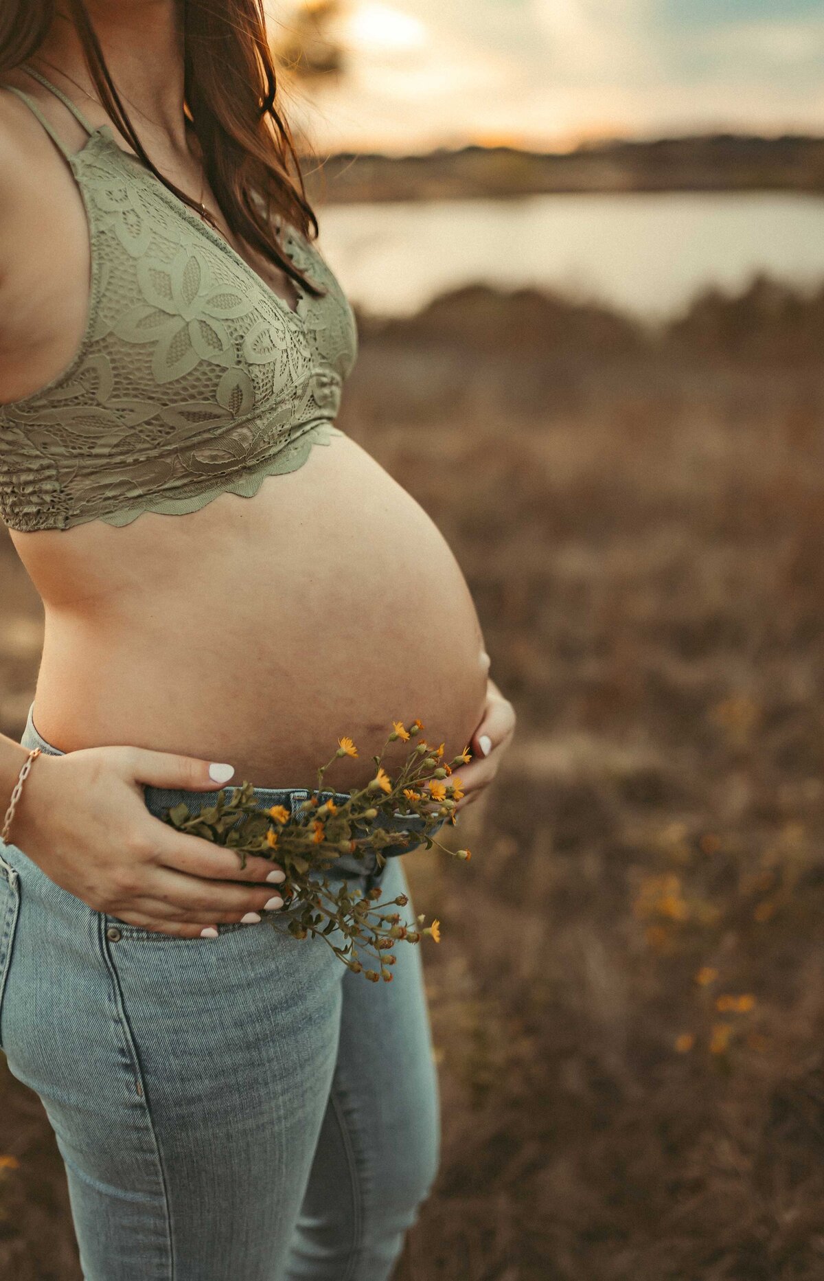 Woman on a maternity session weariing jeans and a lace bralette. She is holding a bunch of small yellow flowers in her hand.