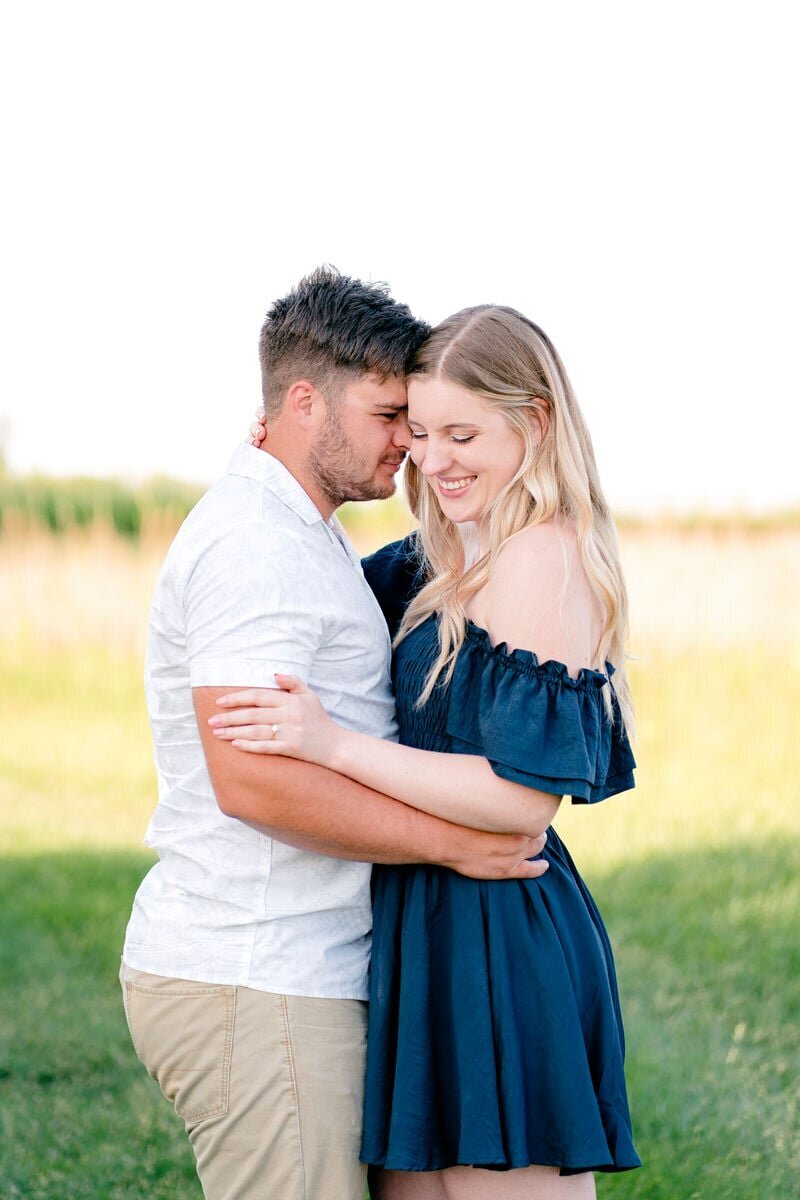A couple embracing outdoors, with the man gently leaning his forehead against the woman's. The woman is wearing a navy off-the-shoulder dress and smiling.